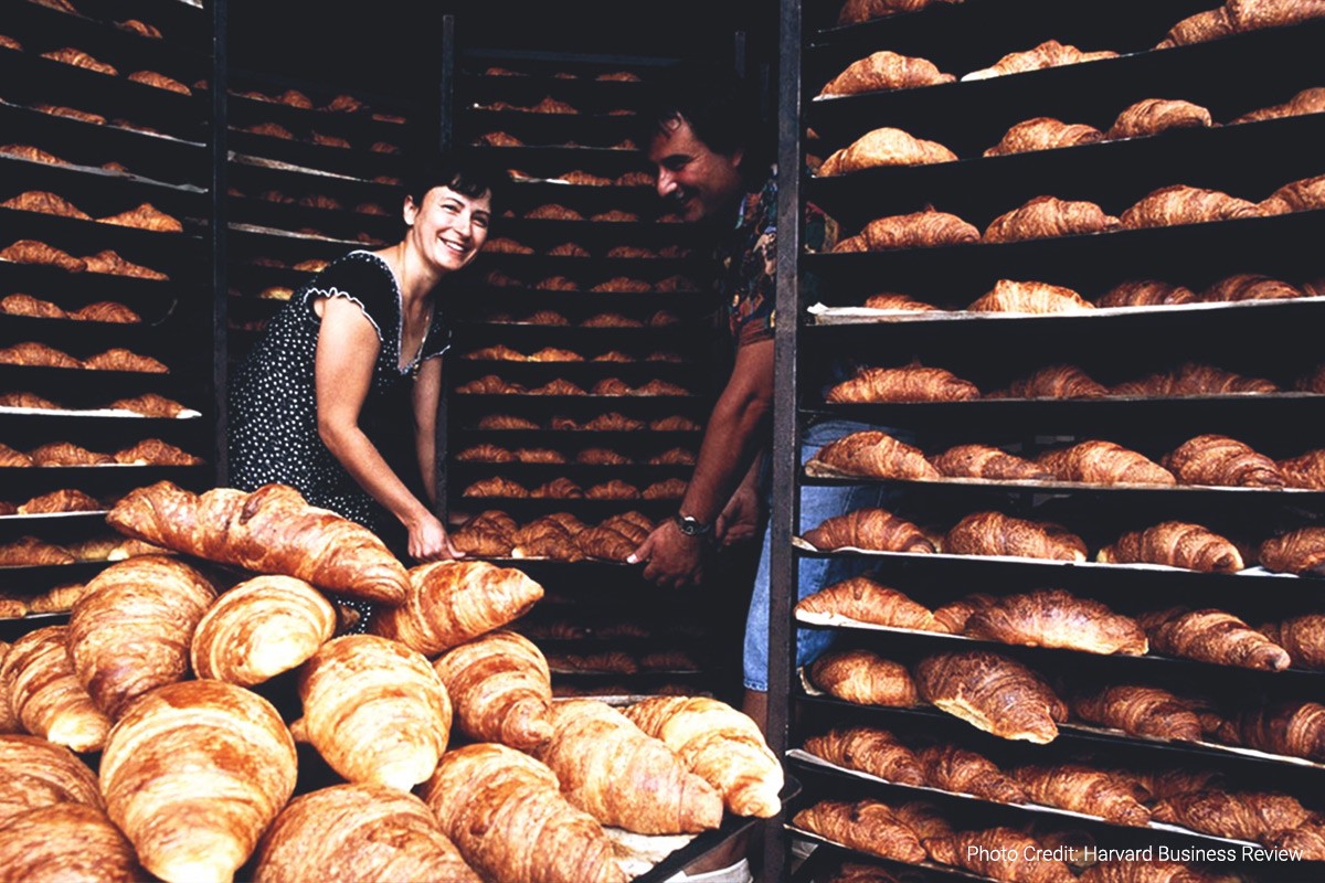 a woman and a man hold a tray while surrounded by racks and racks full of fresh-baked croissants (photo credit: harvard business review)