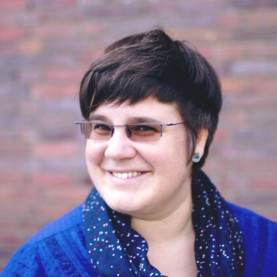 claudia meier, a brunette woman who's wearing a glasses and a blue top, smiles while standing in front of a brick wall