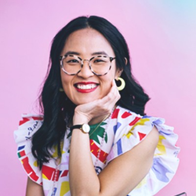 ahran lee, a woman with long dark-brown hair who’s wearing glasses and a multicolored sleeveless top, smiles while sitting in front of a pink-colored background