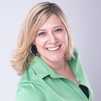 heather ramsey, a woman with shoulder-length blond hair who’s wearing hoop earrings and a green blouse, smiles while sitting in front of an off-white background