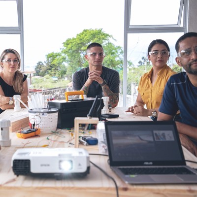 two women and two men sit around a wooden workbench covered in tools and parts while engaging via video with remote team members