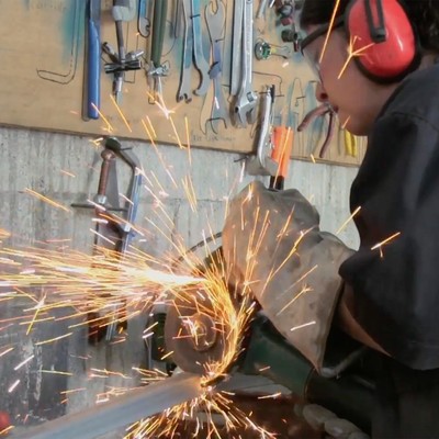 a woman wearing protective gear uses an angle grinder to cut a piece of metal (photo credit: actuality media)