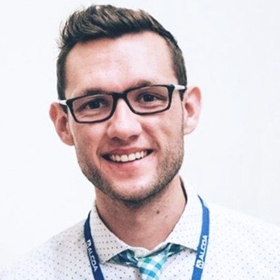 adam stoffel, a man with short brown hair who’s wearing glasses and a button-down shirt and tie, smiles while standing in front of a light-colored background