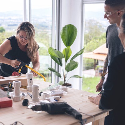 2 women smile and laugh while sawing a piece of wood during a team building activity as colleagues admire their handiwork