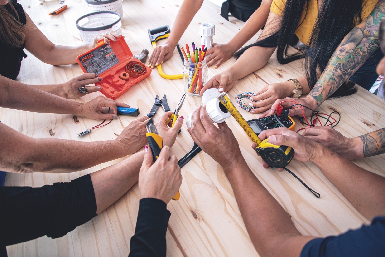 an over-the-shoulder view of the arms and hands of seven onsite workshop participants holding a variety of tools over a wooden workbench