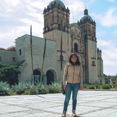 estrella, wearing jeans and a puffy vest, stands smiling in front of santo domingo in oaxaca on a sunny day