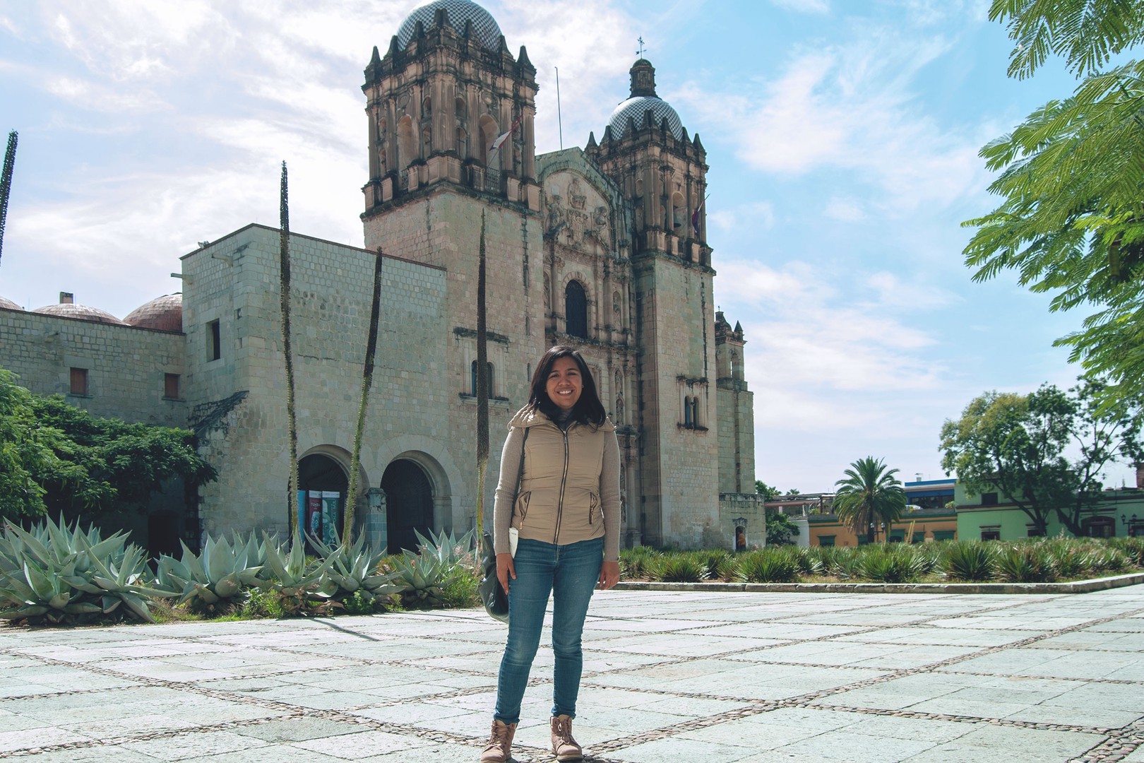 estrella, wearing jeans and a puffy vest, stands smiling in front of santo domingo in oaxaca on a sunny day