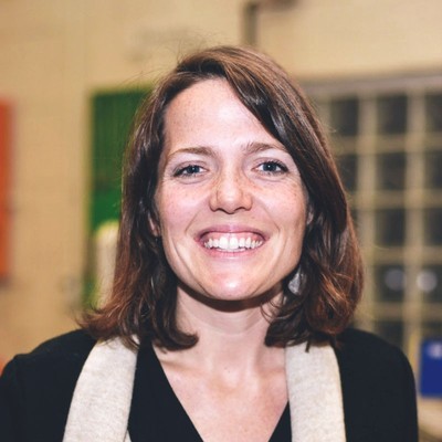 sher vogel, a woman with shoulder-length brown hair who’s wearing a white scarf, smiles while standing in a building