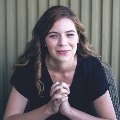 brianna brown, a woman with long brown hair who’s wearing a black short-sleeved top, smiles while leaning forward and clasping her hands