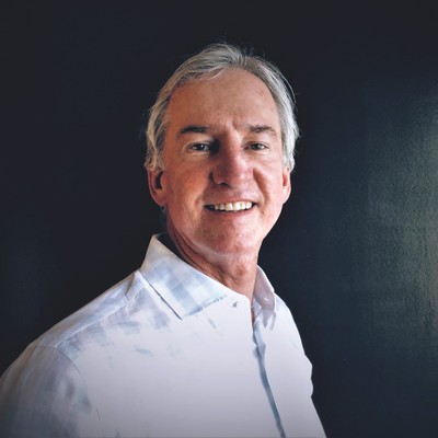 tom malone, a man with short gray hair who’s wearing a white button-down shirt, smiles while standing in front of a dark-colored background