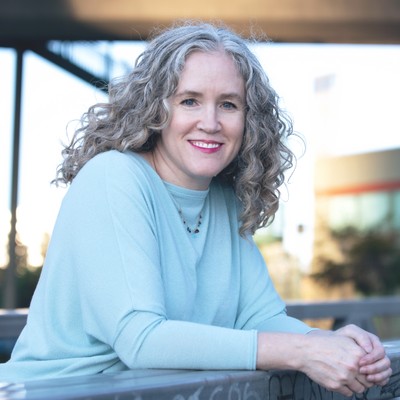 liz hunt, a blond woman with blue eyes who’s wearing a pale blue long sleeve top, leans forward on her arms and smiles