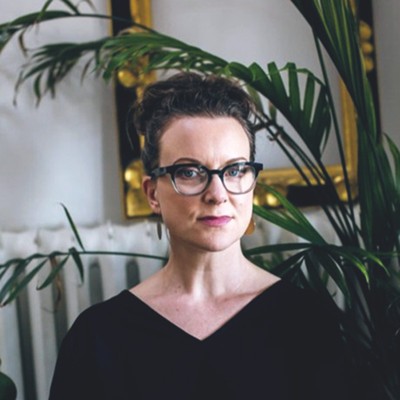 kristy o'leary, a woman with dark-brown bunned hair who’s wearing glasses and a black sweater, smiles while sitting in front of a plant and framed mirror