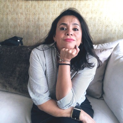 alma vasquez, a woman with long dark-brown hair who’s wearing a blue and white striped blouse, sits on a couch resting her chin on her fist and smiles