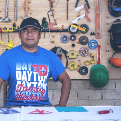enoc, wearing a t-shirt and ballcap, sits in his workshop (a wide variety of tools and helmets hang on a wall in the background)