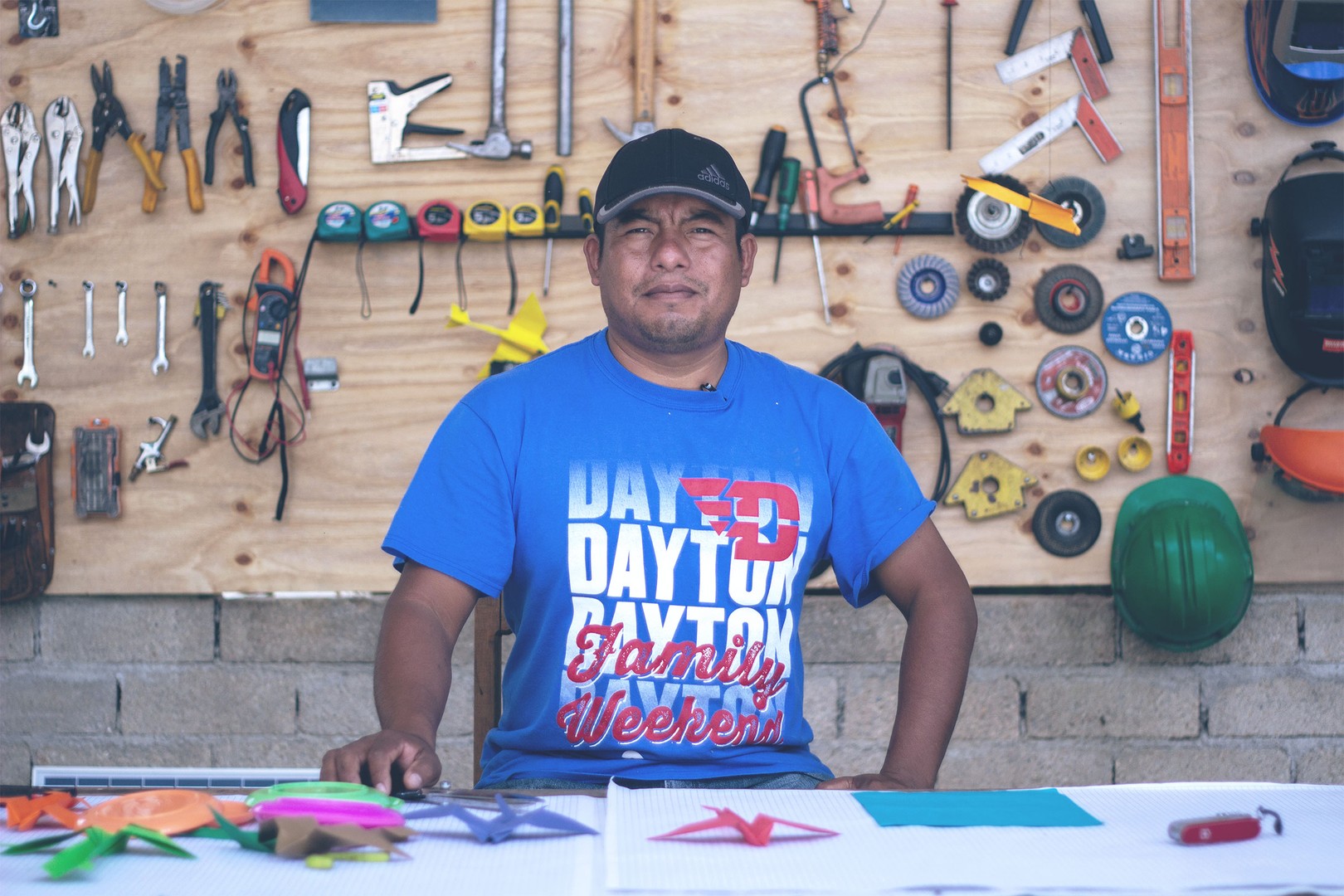 enoc, wearing a t-shirt and ballcap, sits in his workshop (a wide variety of tools and helmets hang on a wall in the background)